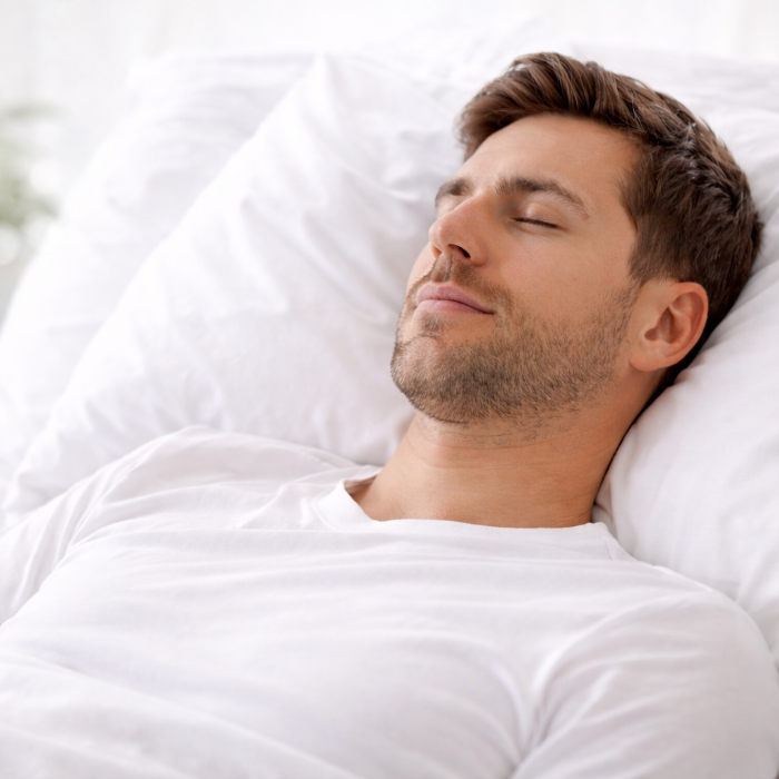 Mann mit 3-Tage-Bart und kurzen braunen Haaren in weißem T-Shirt liegt im Bett und schläft auf einem weißen Kopfkissen